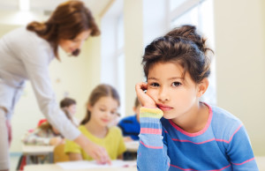 Girl in classroom looks discouraged while teacher helps another student behind her